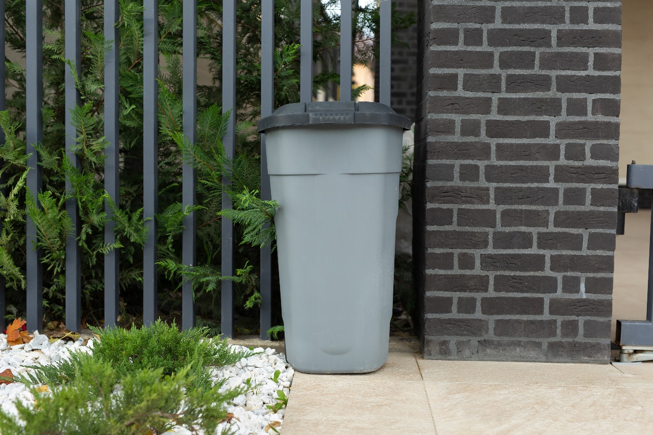 A gray trash bin with a black lid placed next to a black brick wall and a metal fence with green foliage.